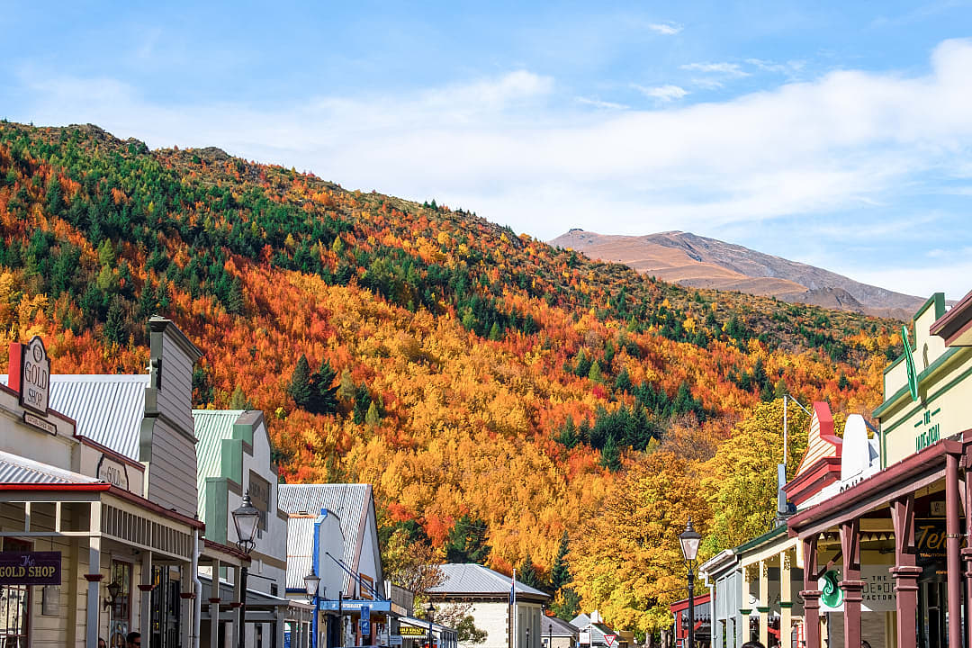Autumn landscape in Arrowtown, New Zealand, with vibrant orange and yellow foliage along the riverbank, surrounded by rugged mountains under a clear sky