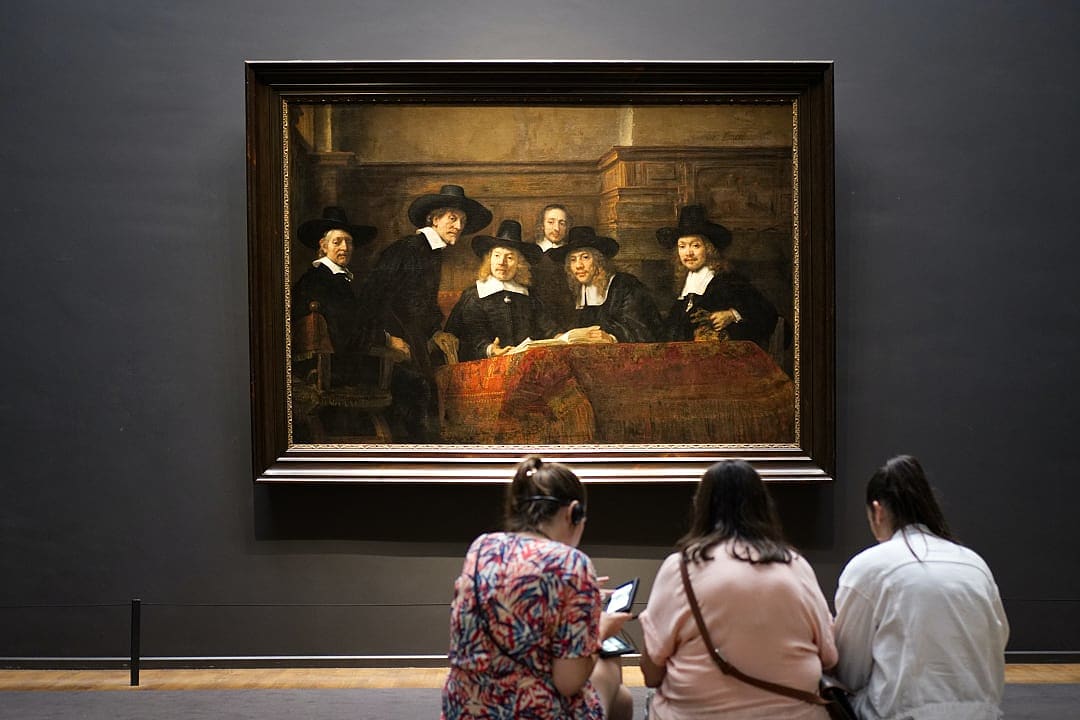 Two women seated on a bench, observing Rembrandt’s group portrait, The Syndics of the Drapers’ Guild, in a gallery.
