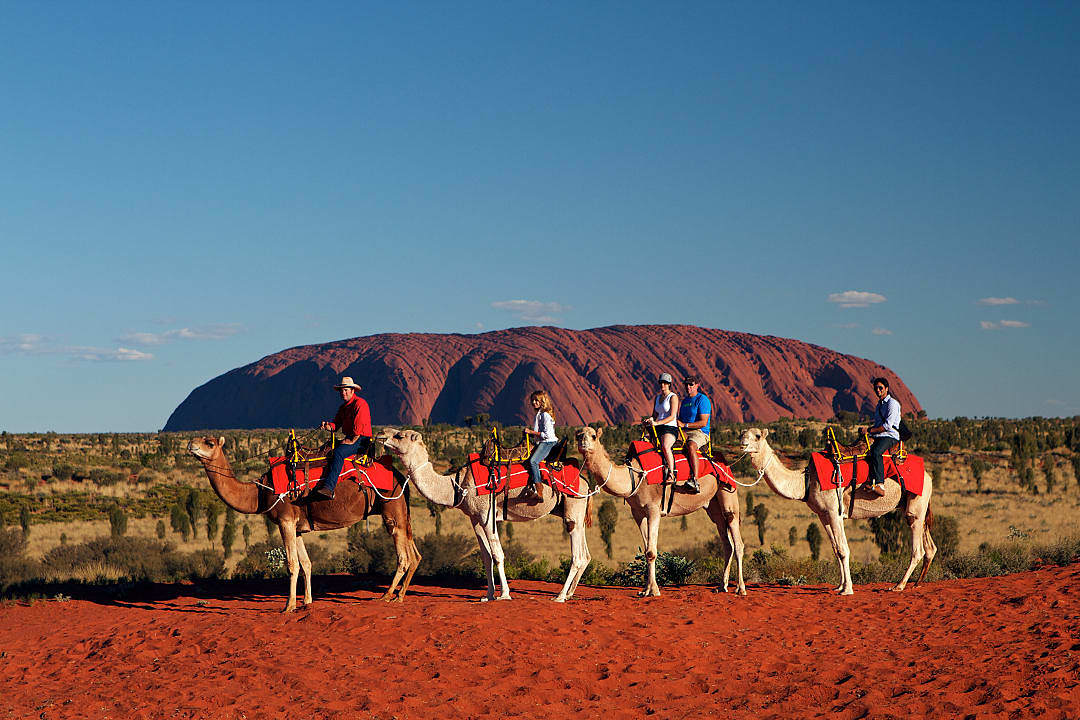 Riders journey across red sands, with Uluru rising behind them.