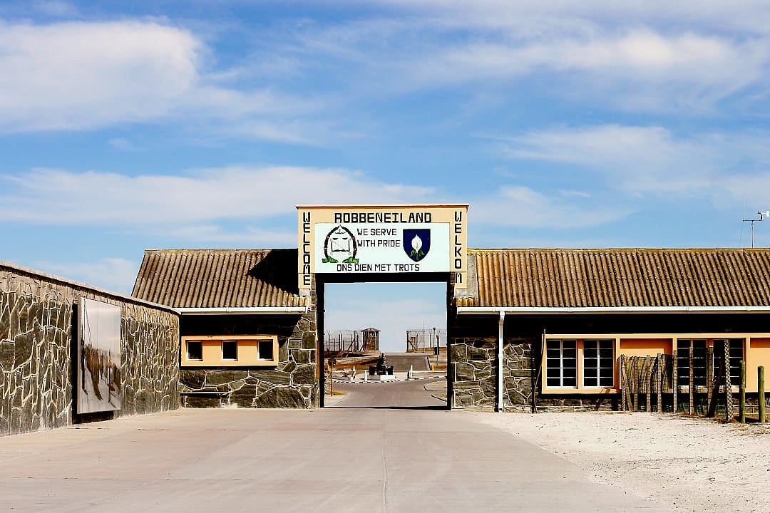 Entrance gate of a historic prison site with stone walls and a welcome sign, symbolizing a place of remembrance and resilience.