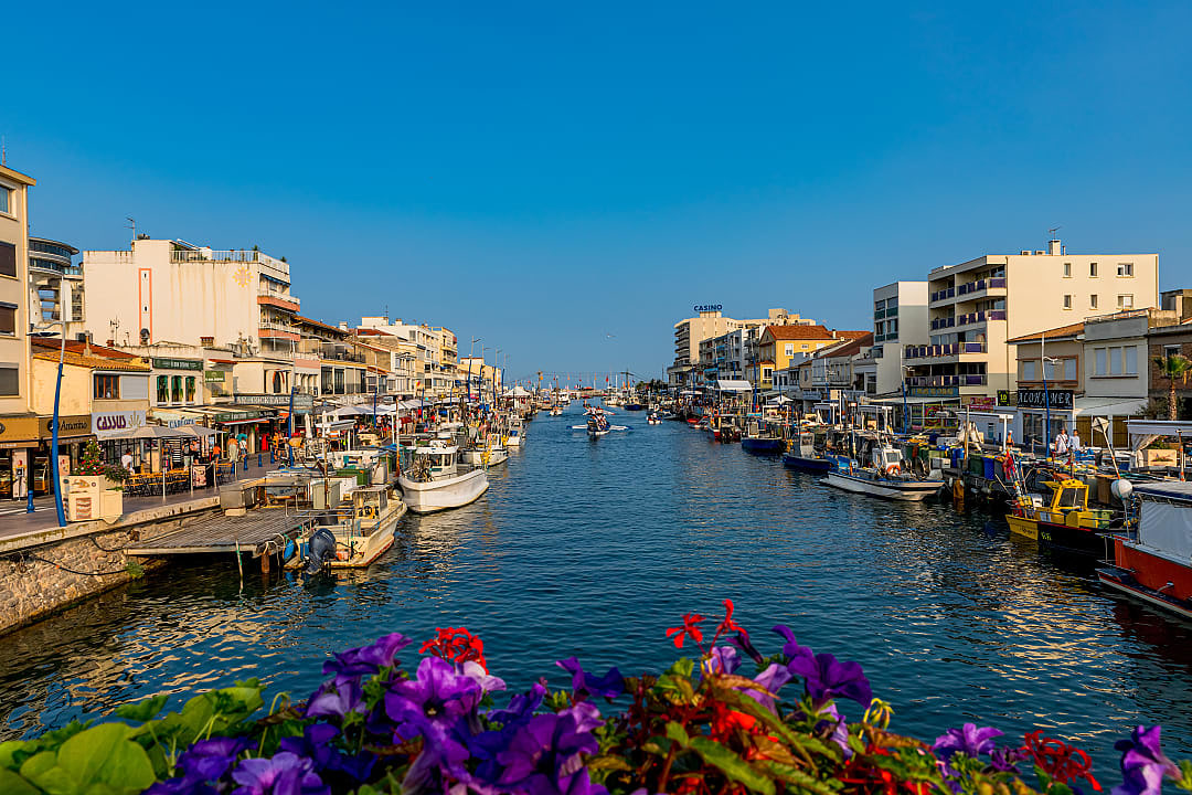 Vibrant view of the River Lez Promenade in Palavas-les-Flots, France, with fishing boats, colorful buildings, and flowers