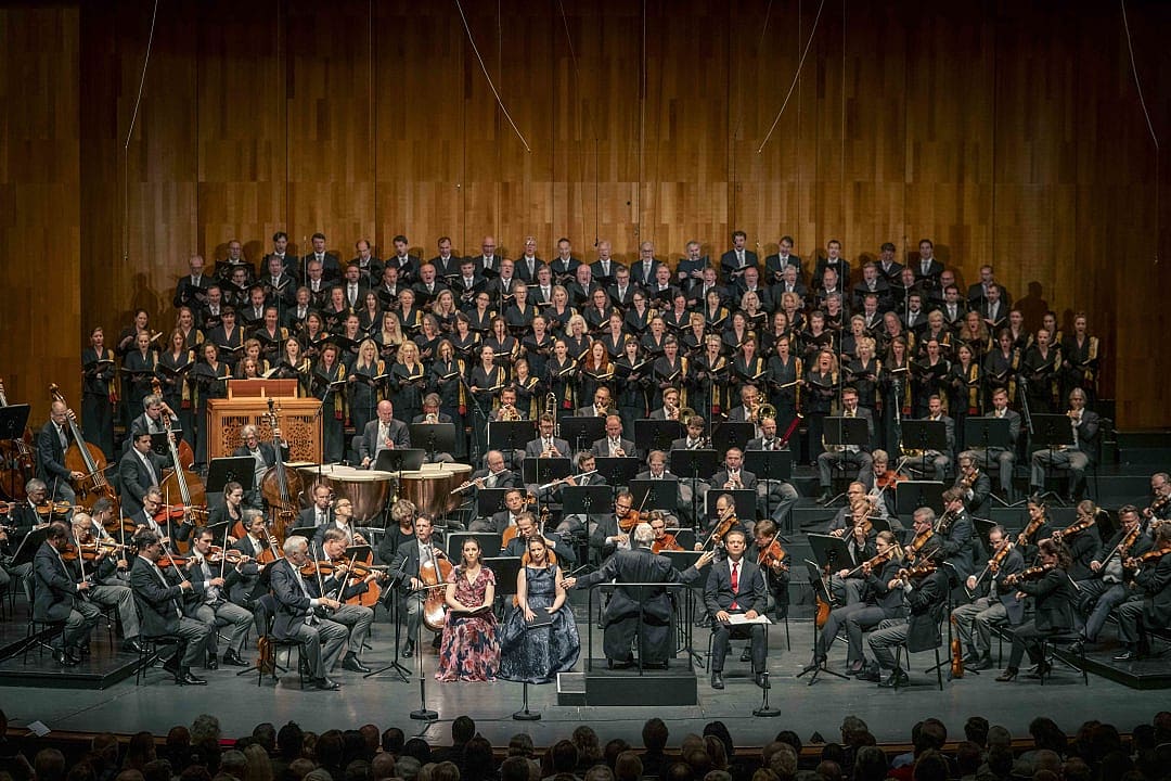 Orchestra and choir performing live on stage with a conductor, soloists, and a wooden backdrop