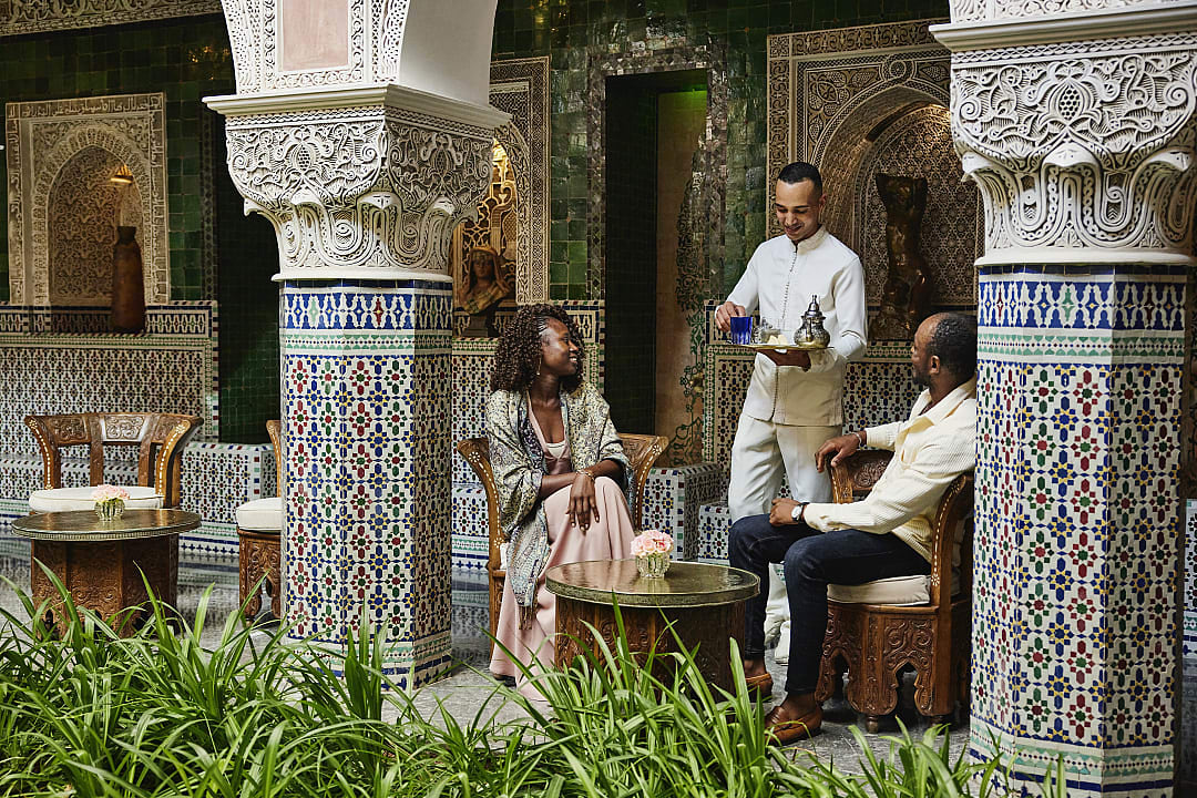 A couple sits in an ornate, tiled courtyard of a traditional Moroccan riad while a staff member serves them.