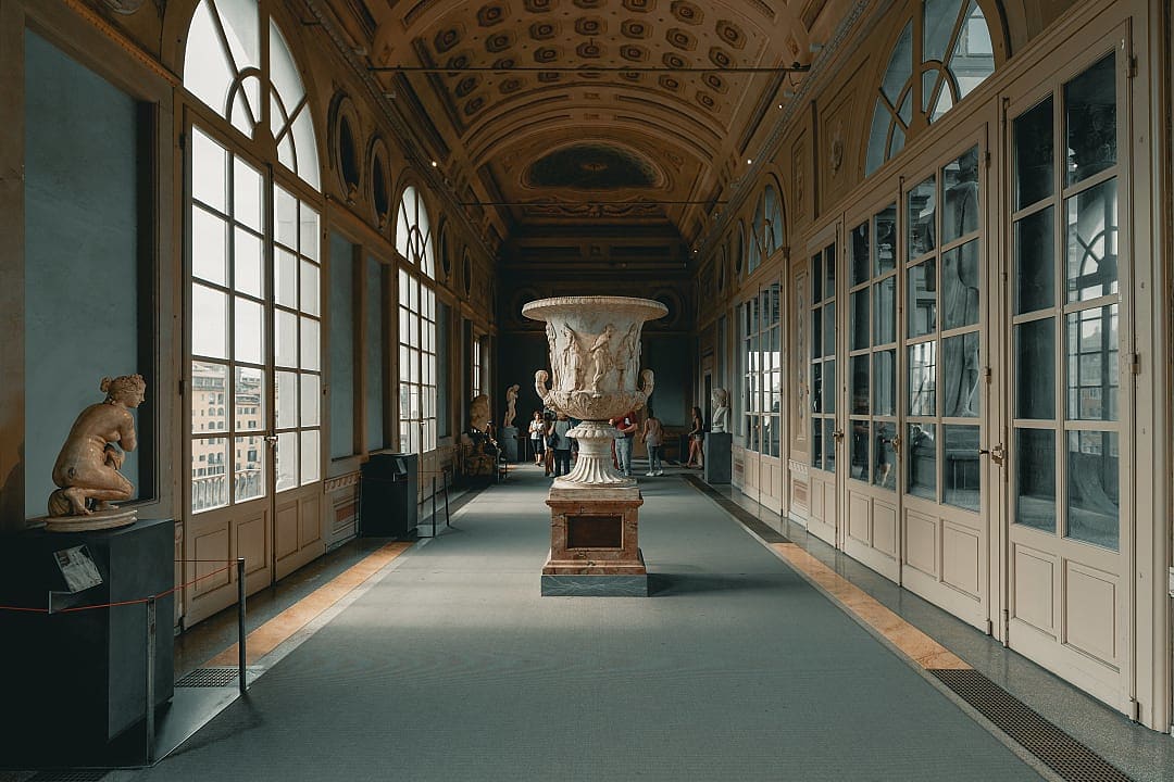 An interior view of a long, sunlit gallery corridor in the Uffizi Gallery, Florence, with classical statues and large windows.