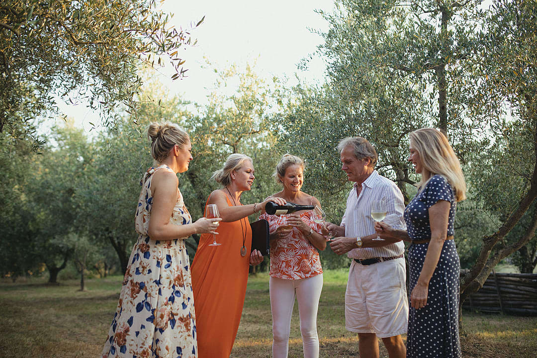 A group of friends stands in a sunny Italian olive grove or vineyard, smiling and talking while holding glasses of wine.
