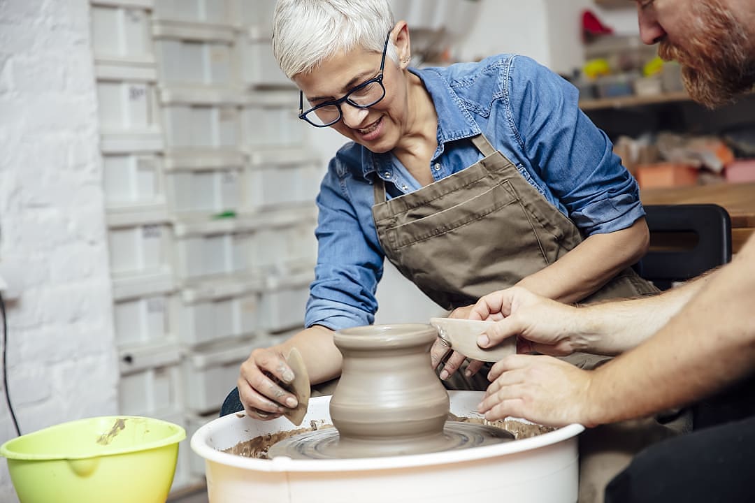 A senior woman with glasses smiling while working with a pottery wheel alongside an instructor in a ceramics workshop.