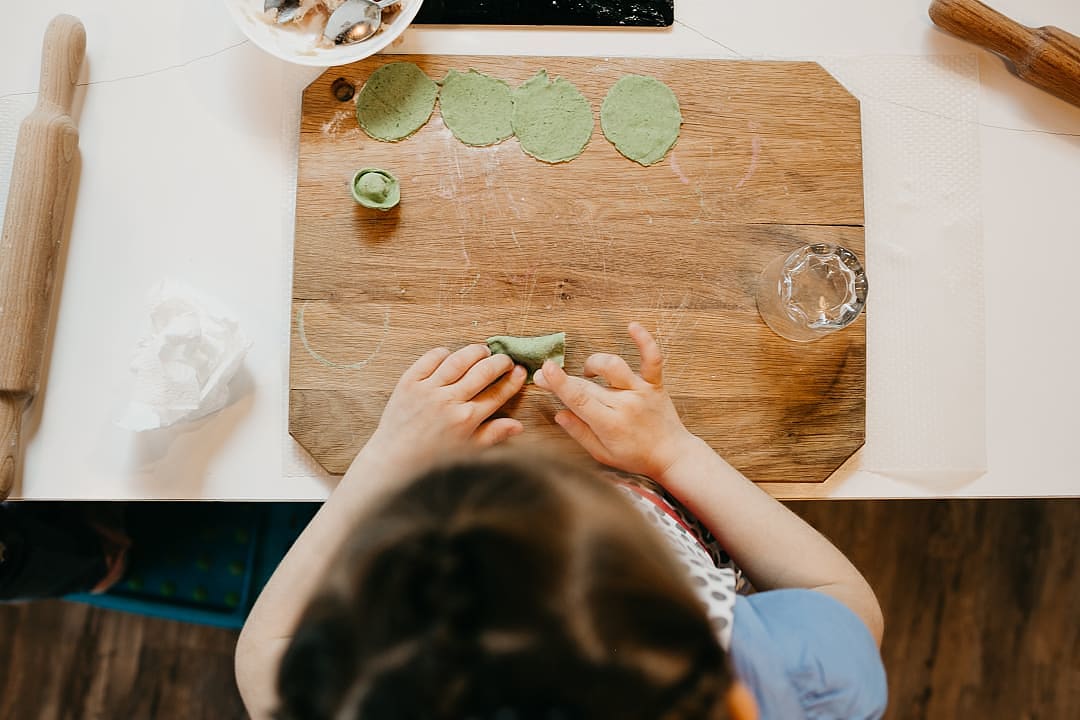A high-angle view of a young child's hands carefully shaping green pasta dough on a wooden board.