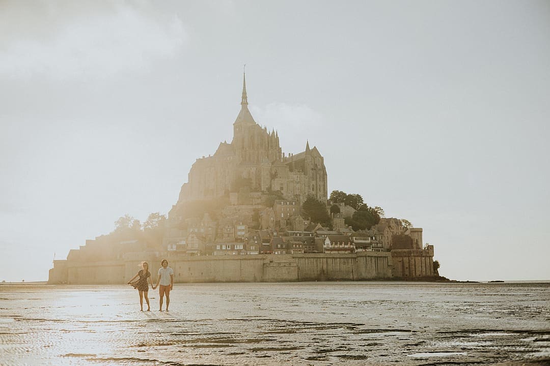 Couple at Mont Saint-Michel in Normandy, France, perched on a rocky island surrounded by tidal waters, showcasing medieval architecture and a dramatic skyline against a cloudy sky