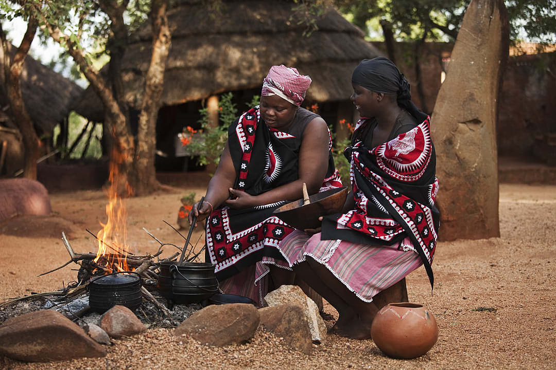 Two women in traditional dress cooking over open fire in a cultural village setting in South Africa