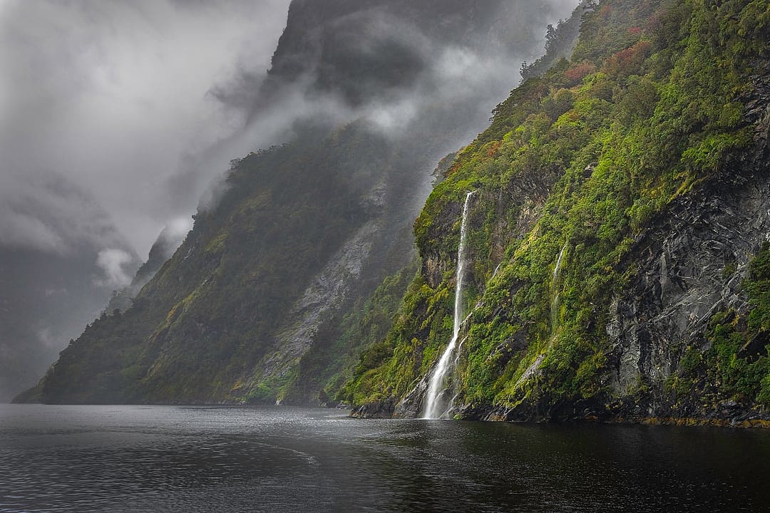 Misty cliffs and waterfalls plunge into peaceful Doubtful Sound waters.