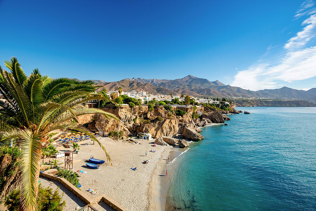 Beach in Nerja, Spain, with palm trees, crystal-clear water, and rocky cliffs in the background."  Let me know if you'd like any changes