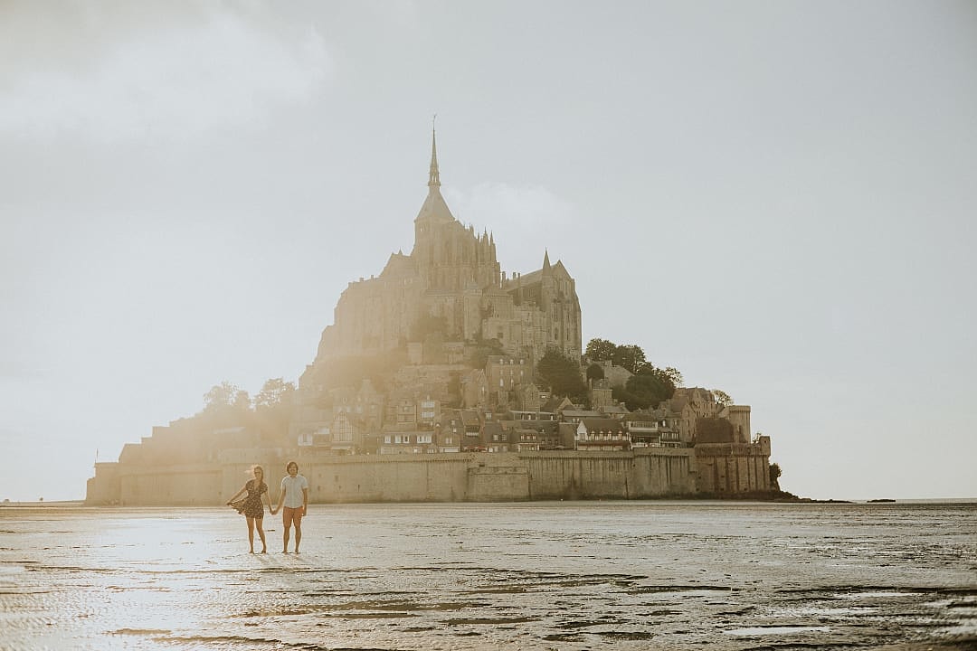 Couple at Mont Saint-Michel in Normandy, France, perched on a rocky island surrounded by tidal waters, showcasing medieval architecture and a dramatic skyline against a cloudy sky