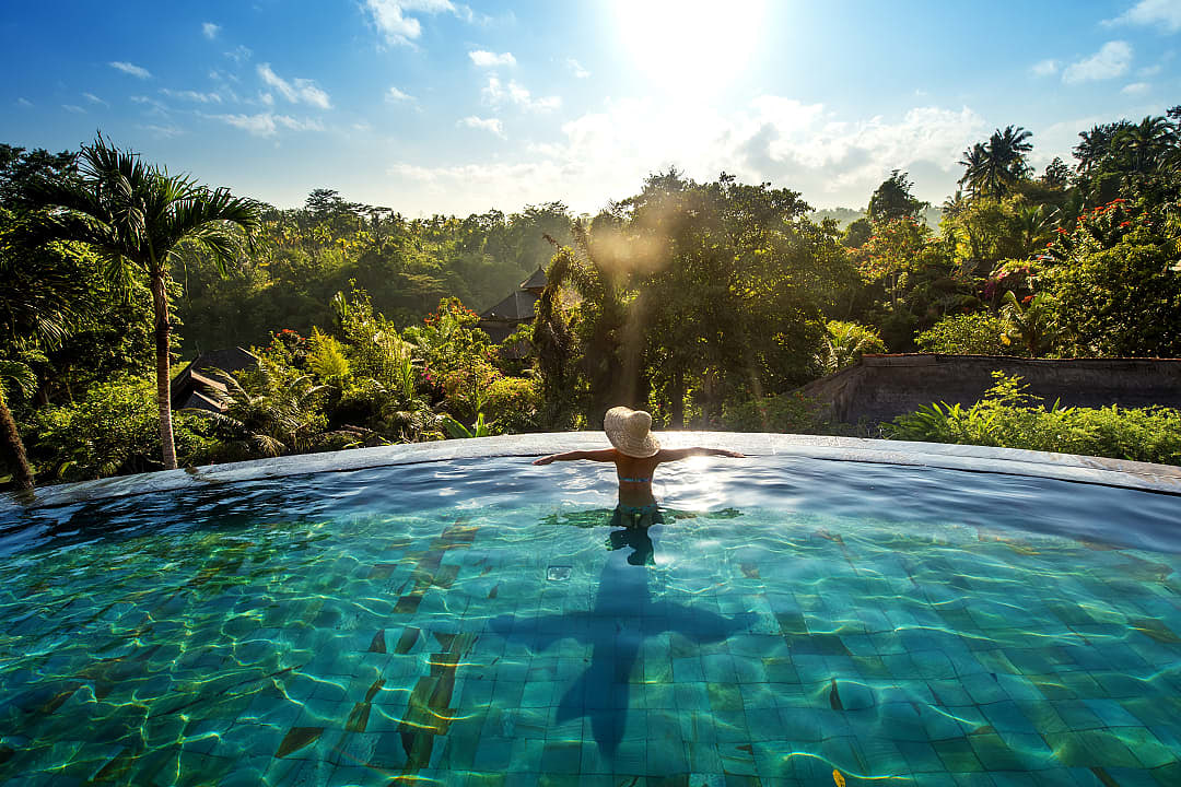 A woman enjoying the sunlight and forest view from a pool in Bali.
