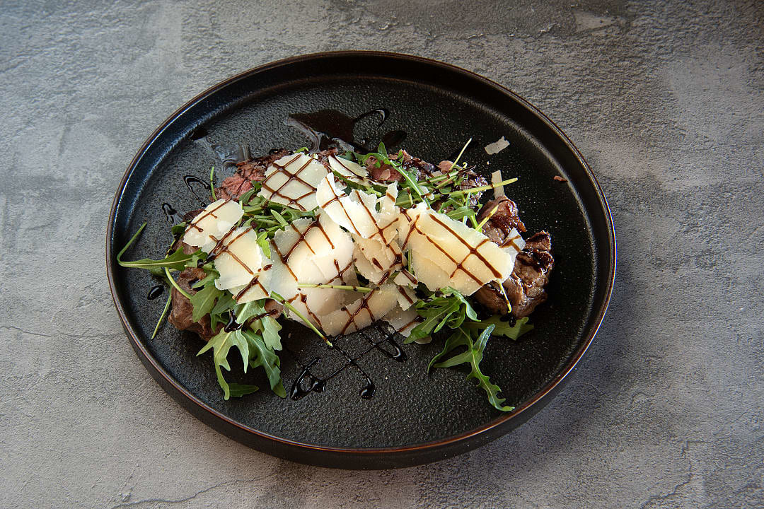 Tagliata di Manzo, Grilled steak topped with arugula, shaved Parmesan, and balsamic glaze, on a black plate