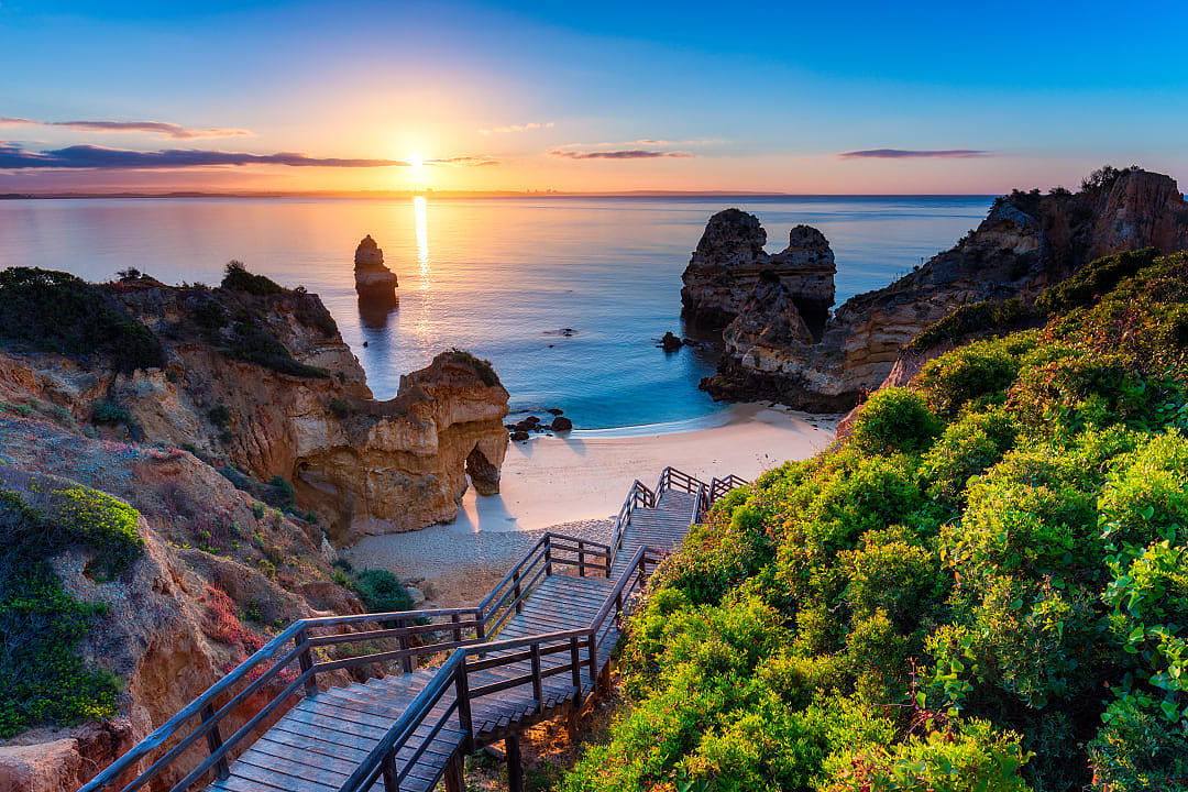 Wooden footbridge to beach Praia do Camilo with the sun setting on horizon of the tranquil blue ocean in the Algarve, Portugal