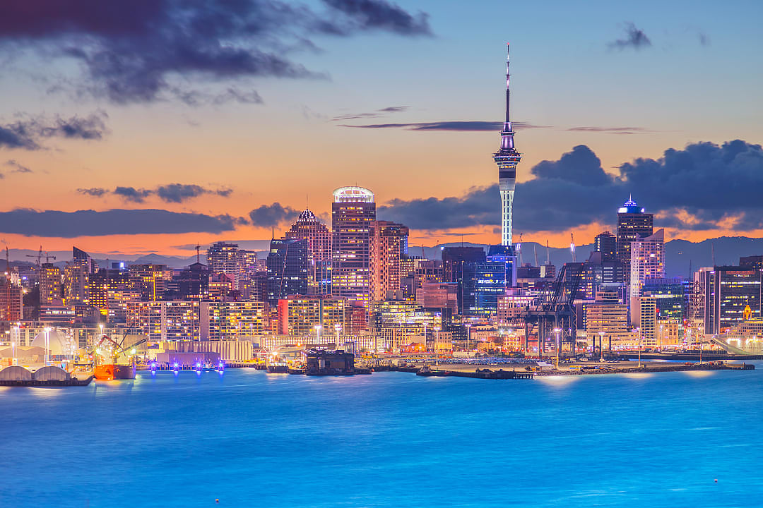 Scenic view of Auckland city skyline at dusk with Sky Tower illuminated, reflecting over the harbor, under a vibrant sunset sky