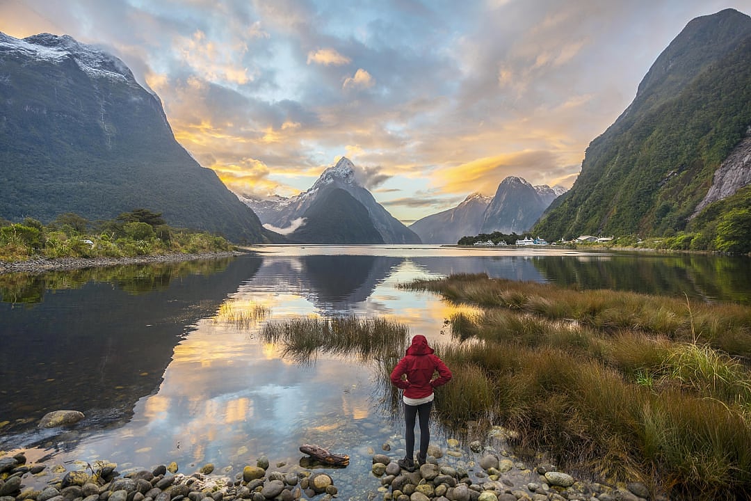 Traveler stands at fjord’s edge under vibrant alpine sunset sky.