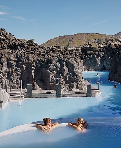 Two women relax in geothermal lagoon surrounded by rocky cliffs.