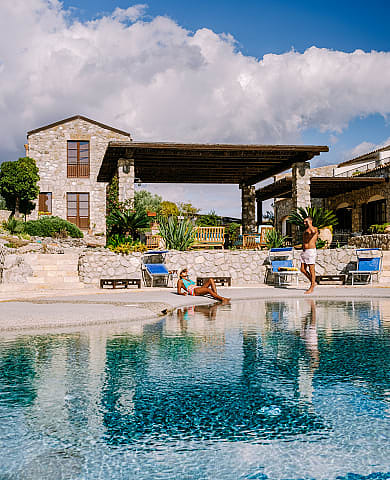 Couple relaxes by stone villa pool under bright blue sky.