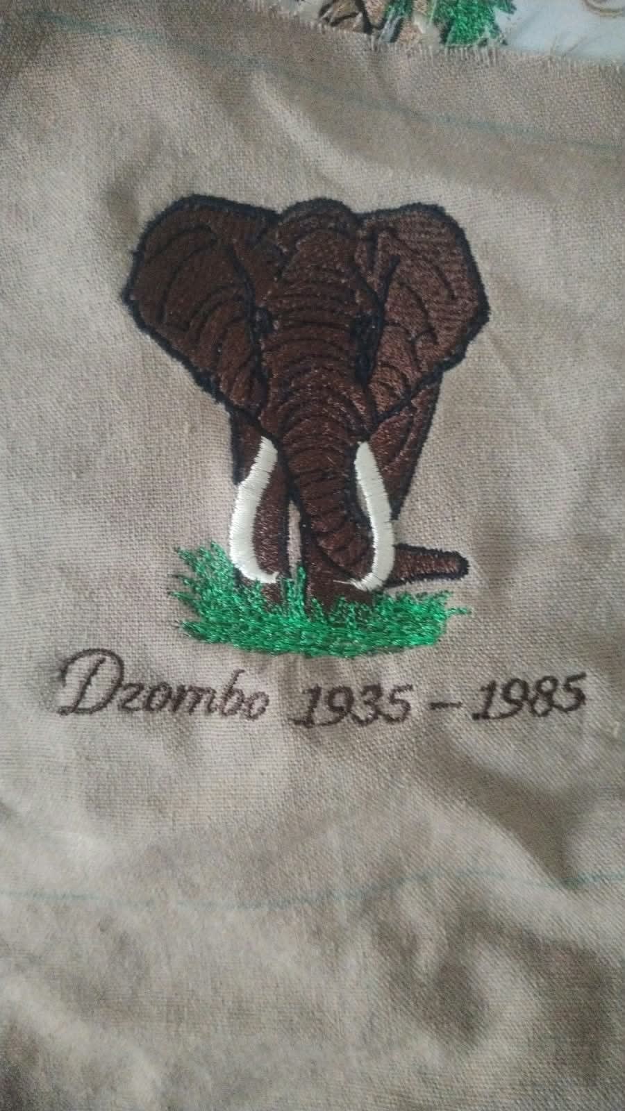 An embroidered brown elephant with white tusks standing in green grass on khaki fabric, above the words Dzombo 1935 - 1985 in script.
