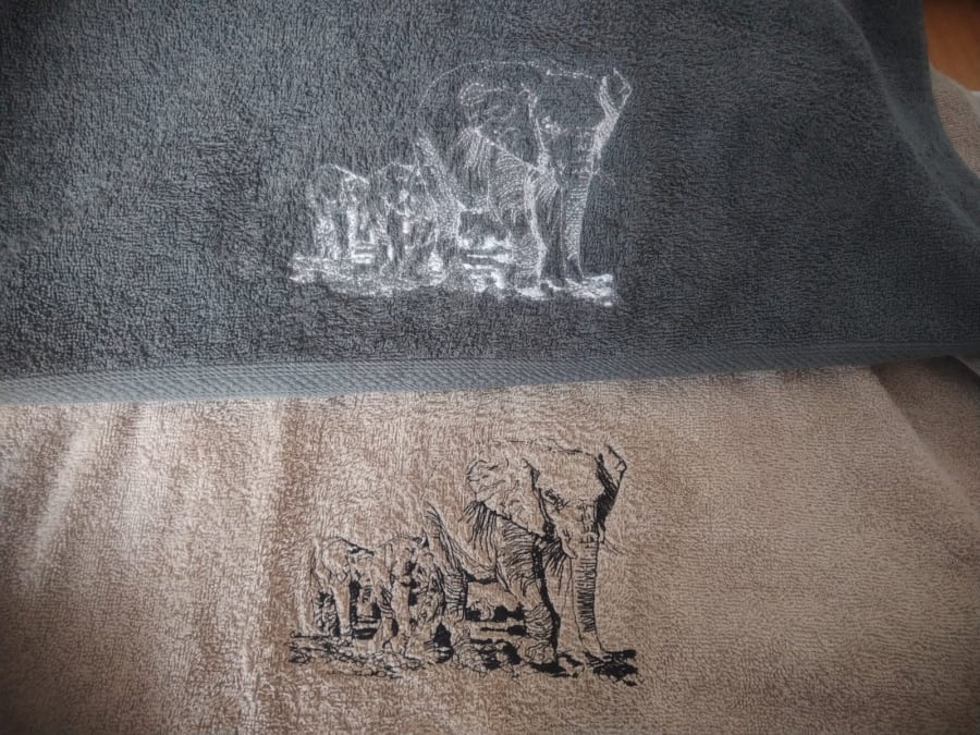 Two towels, one charcoal and one stone-coloured, each embroidered with the same sketch-style elephant and calves, in white thread on the charcoal towel and black thread on the stone towel.
