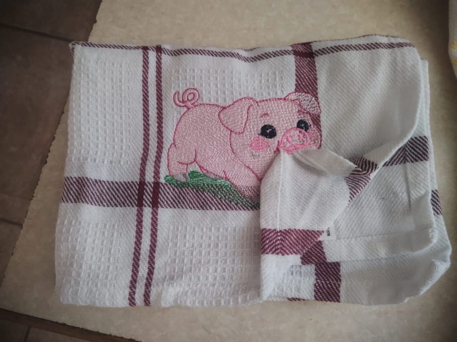 A pink cartoon pig with rosy cheeks standing on a patch of green grass, embroidered on a white tea towel with maroon checks.
