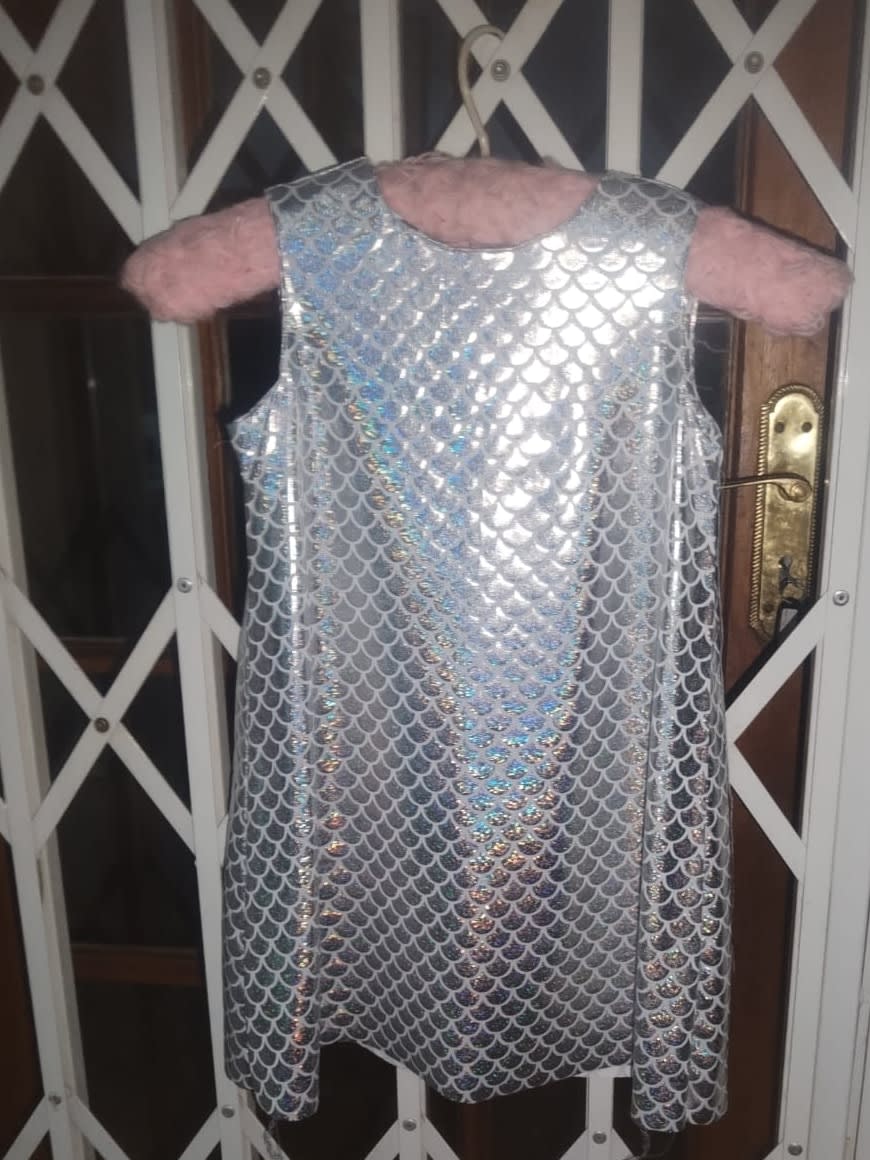 A sleeveless A-line toddler dress in holographic silver mermaid-scale fabric that shimmers pink and blue, hanging on a pink fluffy hanger.