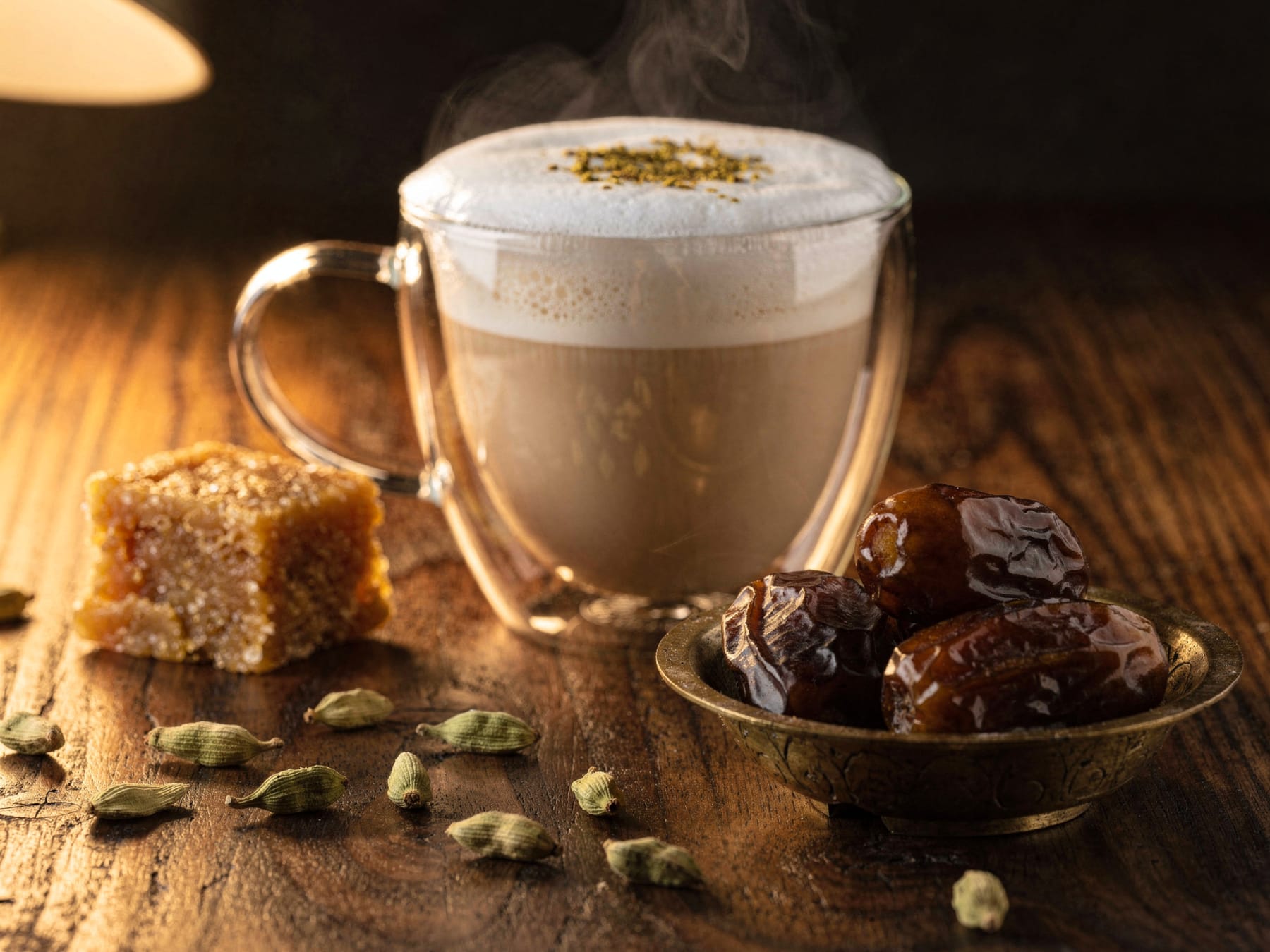A steaming cardamom latte in a glass cup on a dark wooden table, with a block of jaggery, green cardamom pods and a brass bowl of dates