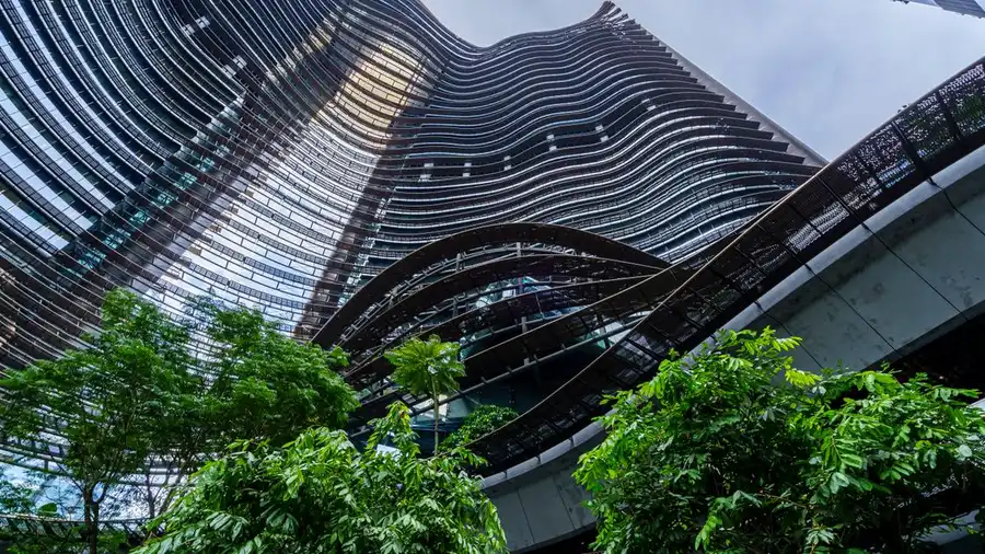 Desain arsitektur bergelombang dari gedung pencakar langit kaca modern di tengah hijaunya tanaman.