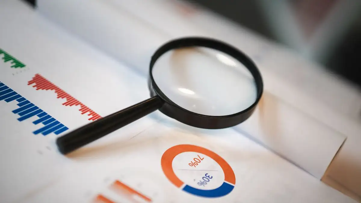 Gambar yang menampilkan kaca pembesar yang diletakkan di atas grafik dan diagram keuangan yang menggambarkan data statistik.