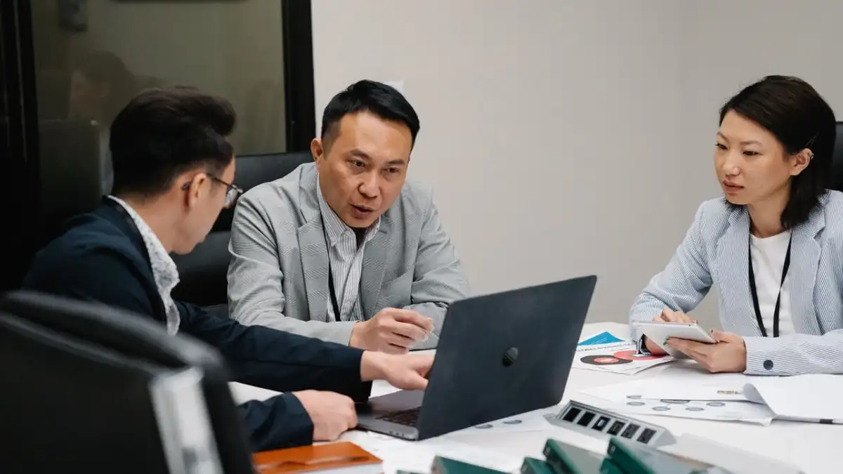Tiga profesional terlibat dalam pertemuan, mendiskusikan strategi di sekitar laptop di lingkungan kantor modern.