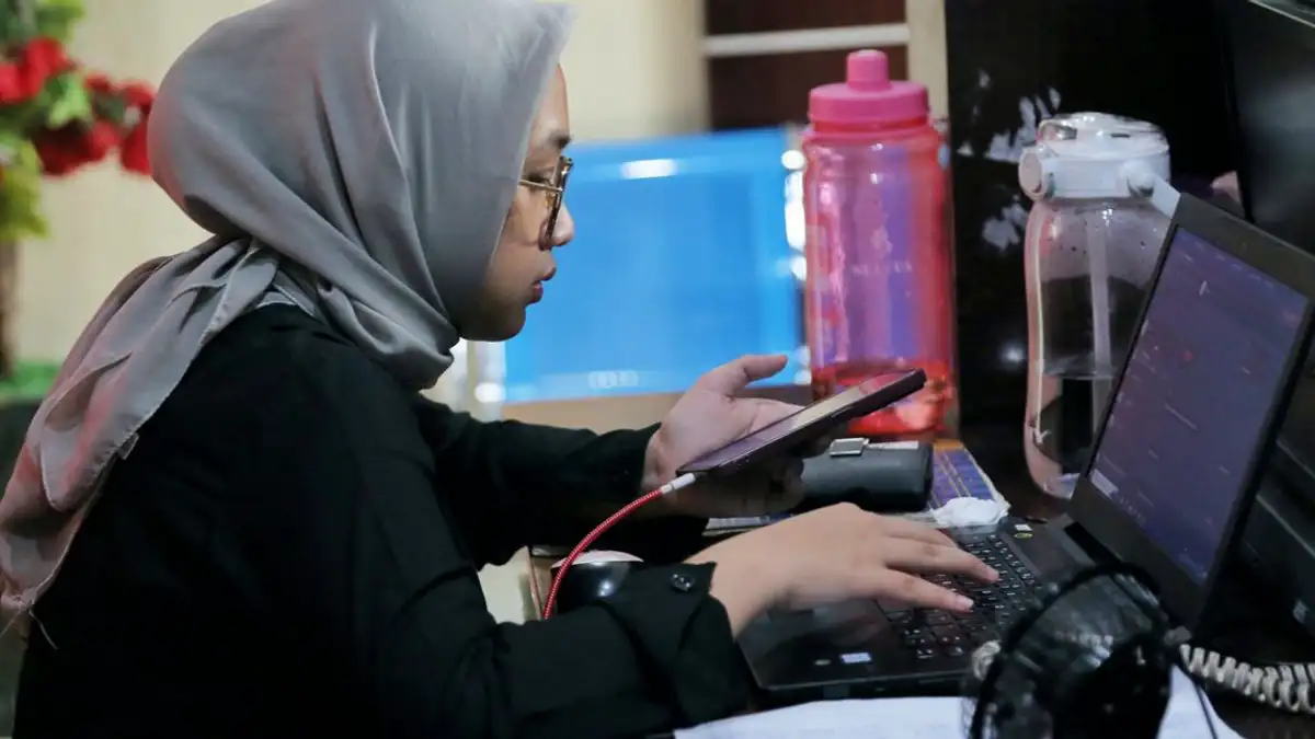 Wanita muda menggunakan telepon dan laptop di kantor, melakukan banyak tugas sekaligus dan fokus pada pekerjaan.
