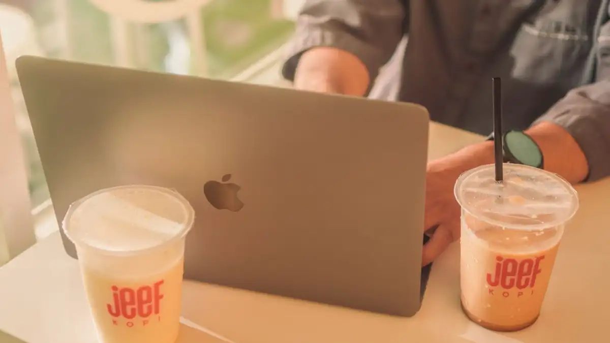 Orang yang bekerja di laptop dengan minuman kopi dingin di kafe, suasana Makassar.
