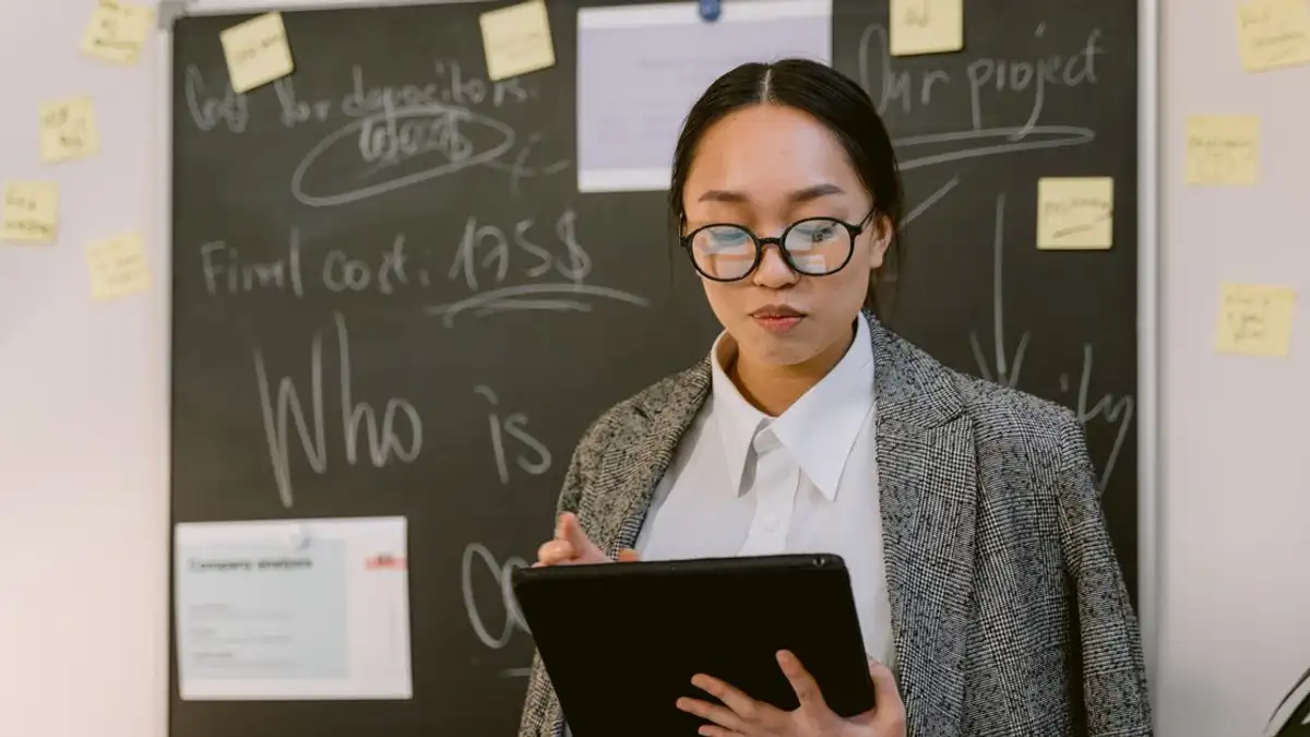 Wanita Asia di kantor yang menggunakan tablet digital, mengenakan kacamata, berdiri di depan papan tulis.