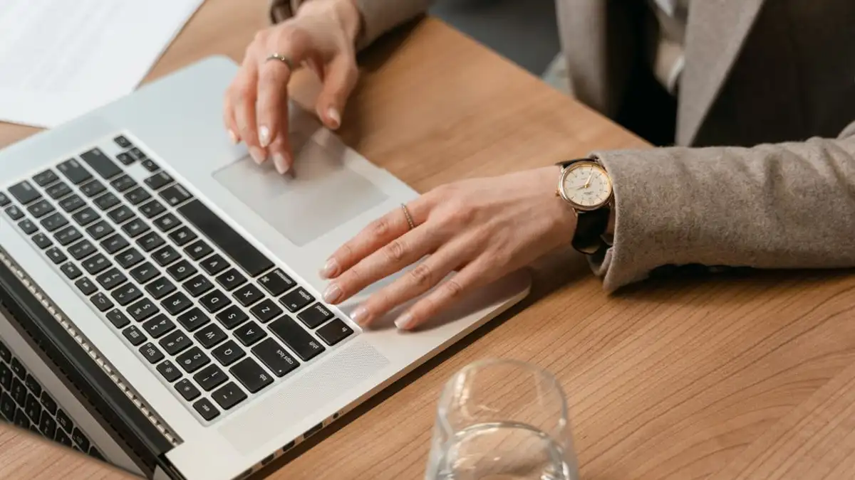 Wanita bekerja di laptop di meja kayu dengan gelas air dan dokumen.