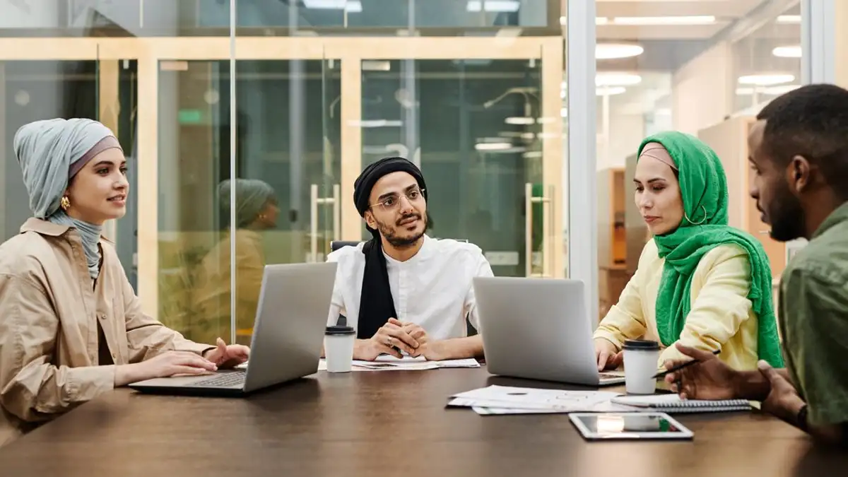 Sekelompok orang yang beragam mendiskusikan ide-ide di lingkungan kantor modern, menggunakan laptop dan tablet.