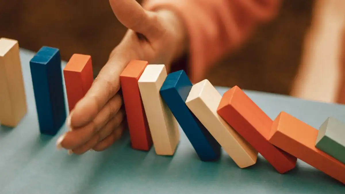 Sebuah tangan secara strategis menghentikan domino biru dan merah yang jatuh di atas meja.