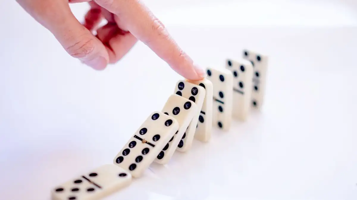 Gambar close-up tangan yang mendorong domino, menggambarkan sebab dan akibat.