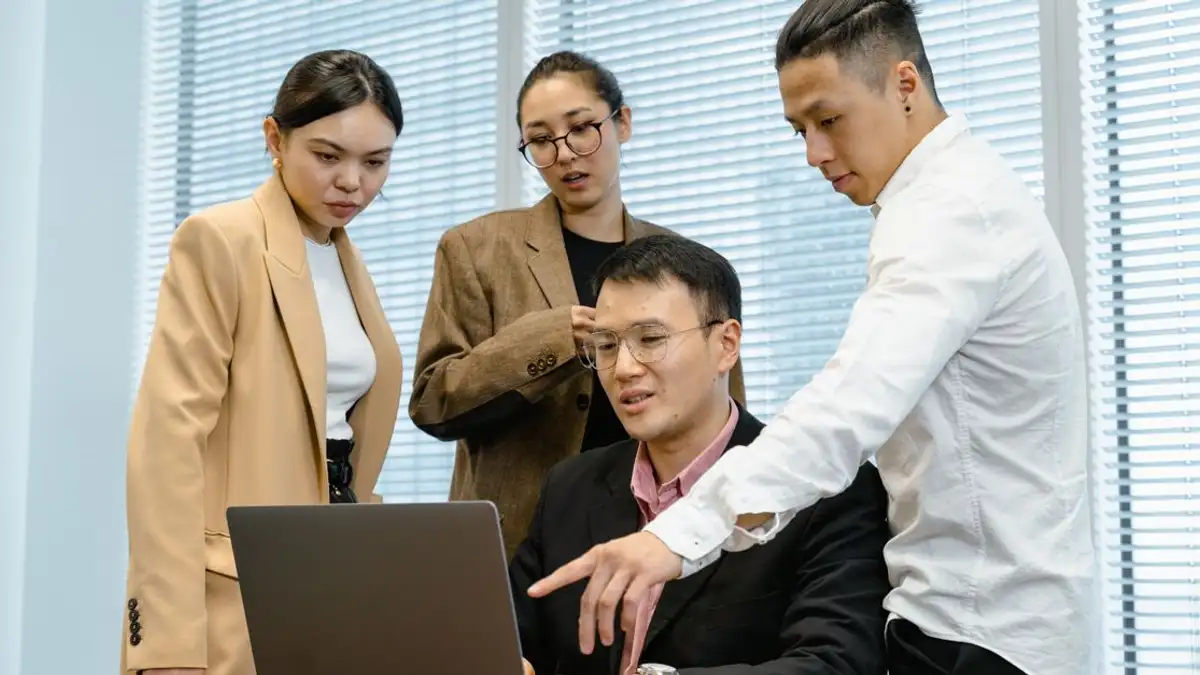 Sekelompok profesional terlibat dalam kerja sama di sekitar laptop di lingkungan kantor modern.