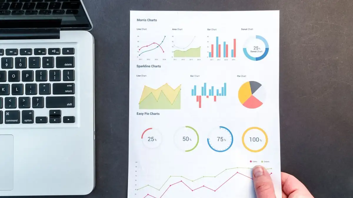 Grafik dan diagram rinci pada dokumen di samping laptop, mewakili analisis data.