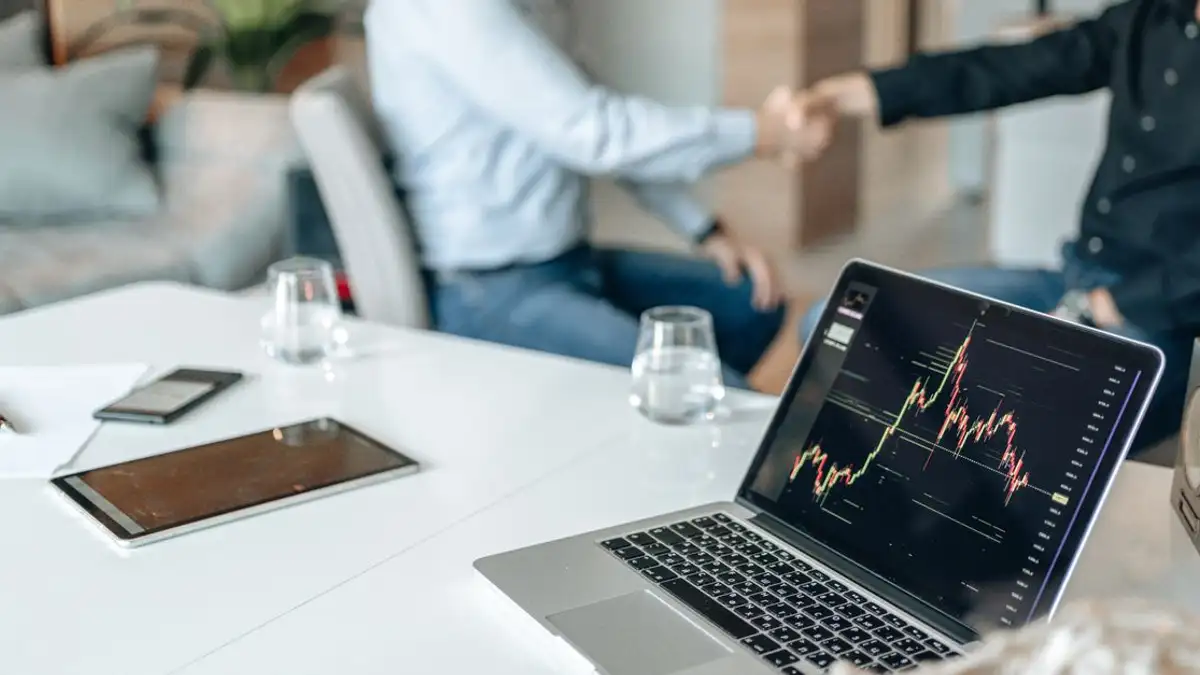 Dua profesional bersalaman di kantor dengan analisis data di layar laptop.