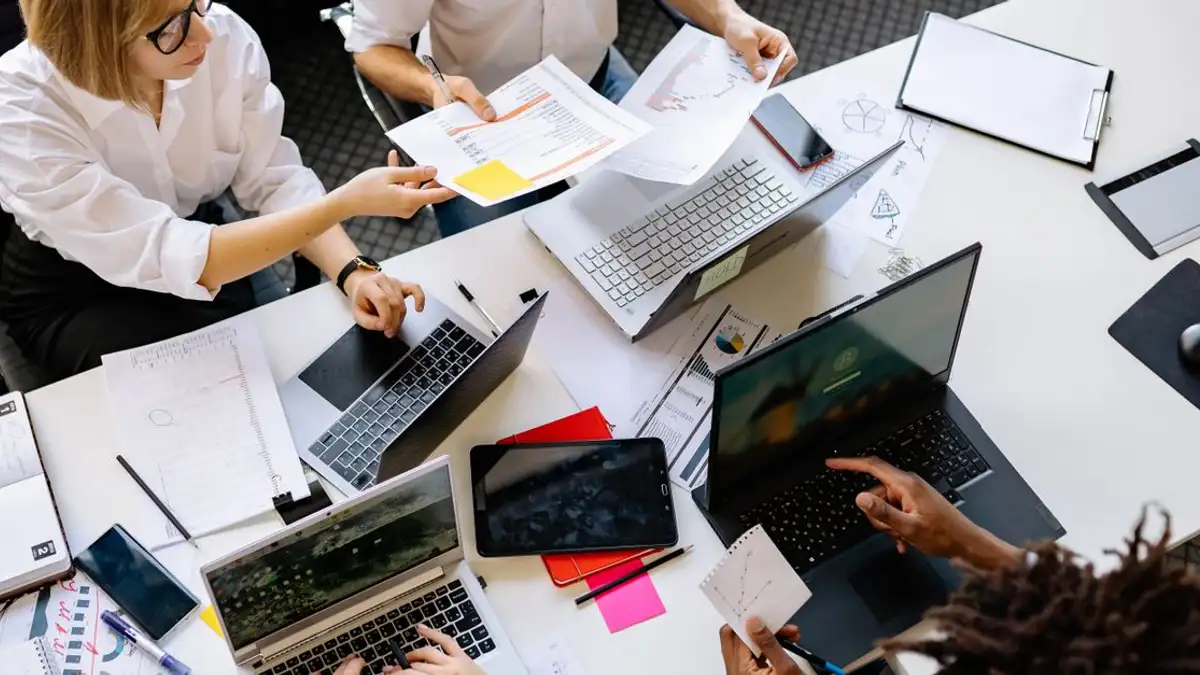 Tampilan atas dari tim kerja yang beragam terlibat dalam pertemuan kantor kolaboratif, mendiskusikan dokumen dan menggunakan laptop.