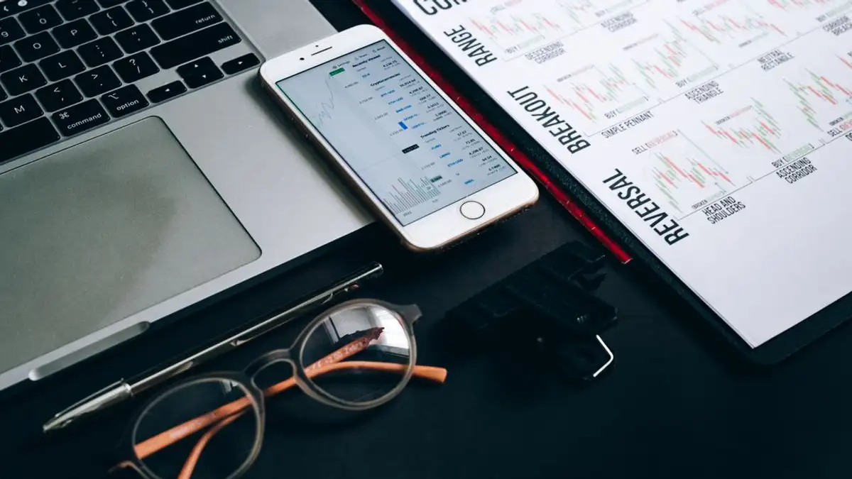 Stasiun kerja dengan laptop, smartphone, kacamata, grafik keuangan, dan pulpen untuk analisis trading.