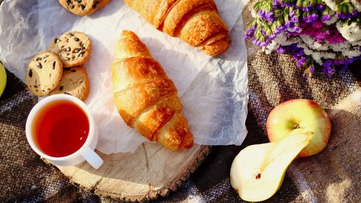 Nikmati sarapan yang nyaman dengan croissant, kue, buah, dan secangkir teh di atas meja yang rustic.