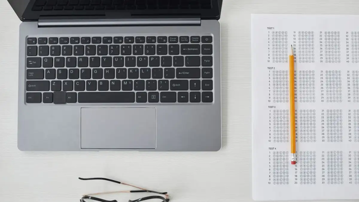 Tampilan datar laptop, lembar ujian, dan pensil di meja putih, sempurna untuk tema pendidikan dan teknologi.