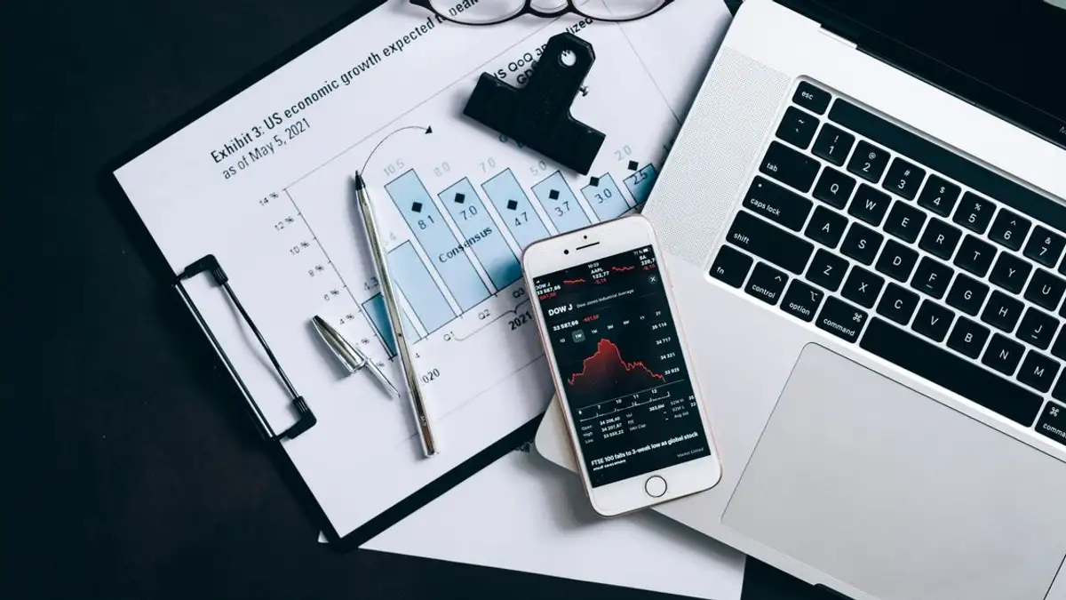 Tampilan atas dokumen keuangan, laptop, dan ponsel yang menampilkan data pasar saham.