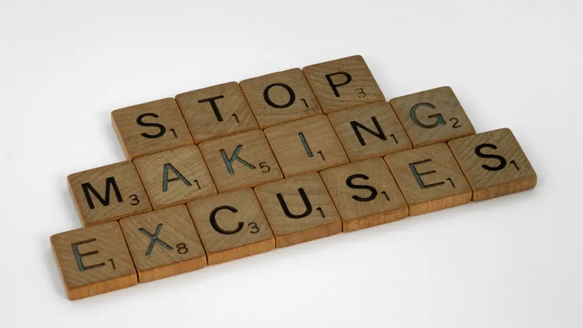 Papan Scrabble kayu mengeja 'Hentikan alasan' di latar belakang putih, mendorong perbaikan diri.