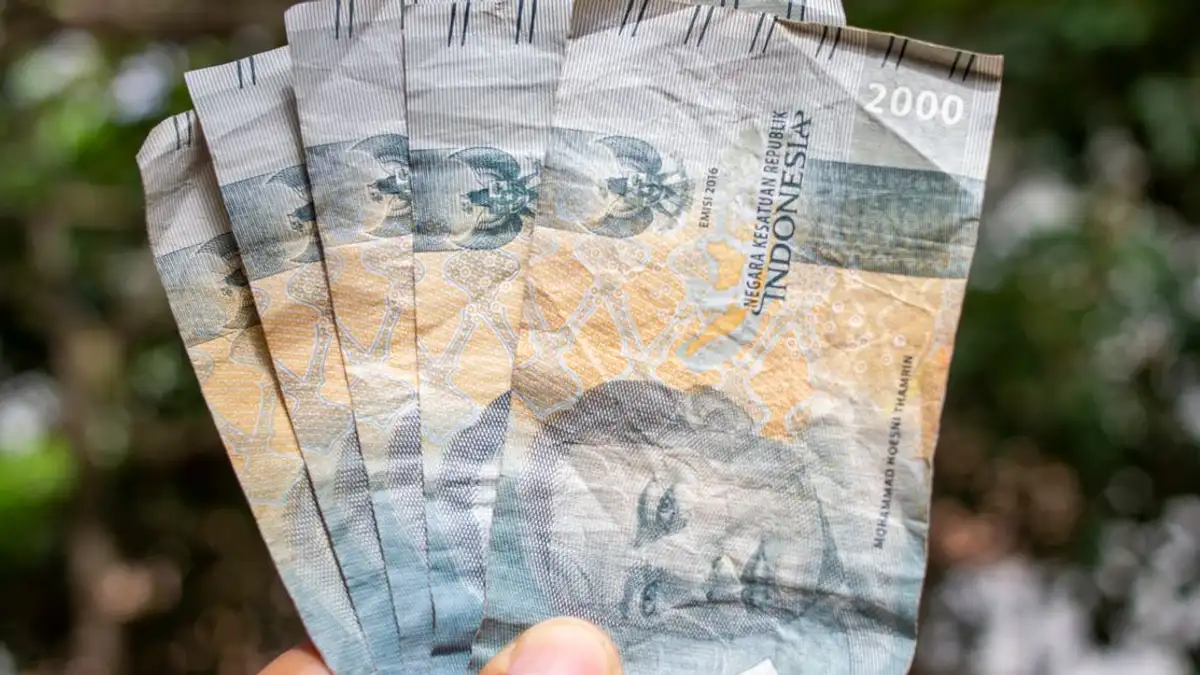 Tangan memegang beberapa uang kertas rupiah Indonesia di luar ruangan, menunjukkan konsep valuta dan keuangan.