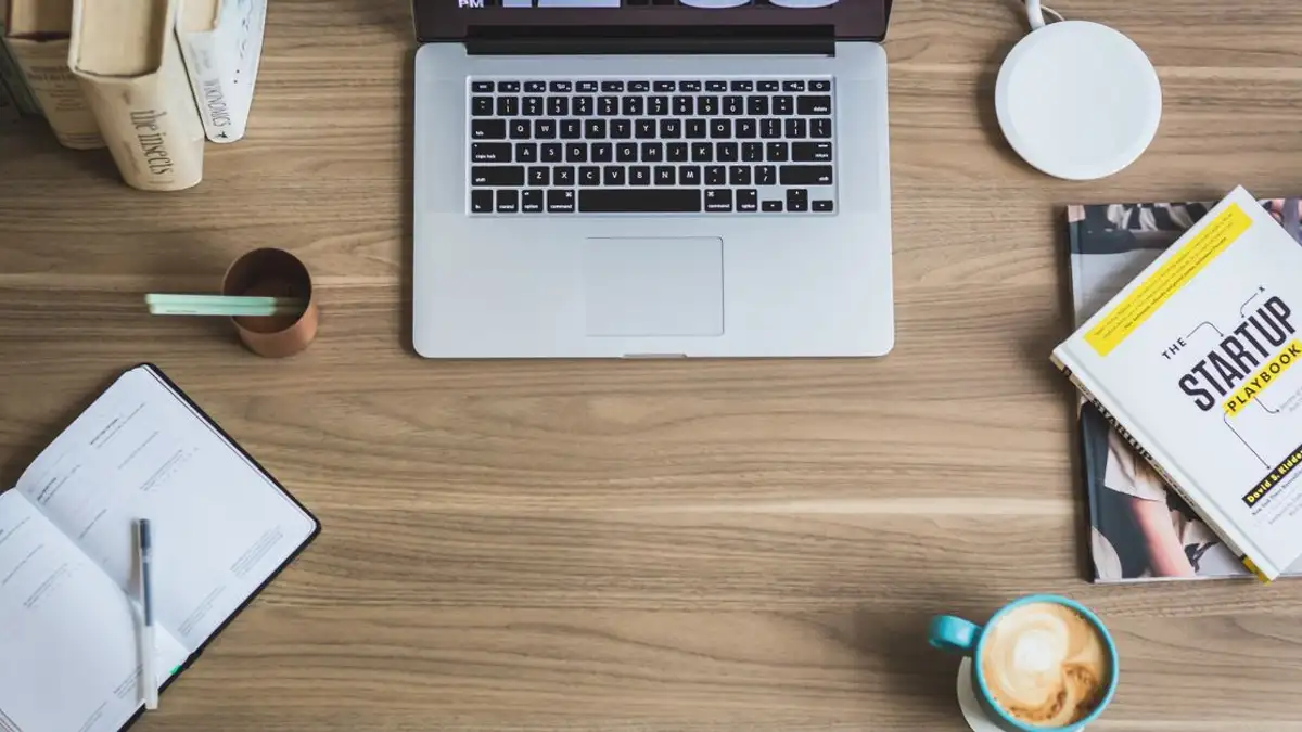 Tampilan datar dari ruang kerja modern dengan laptop, cangkir kopi, dan buku startup di meja kayu.