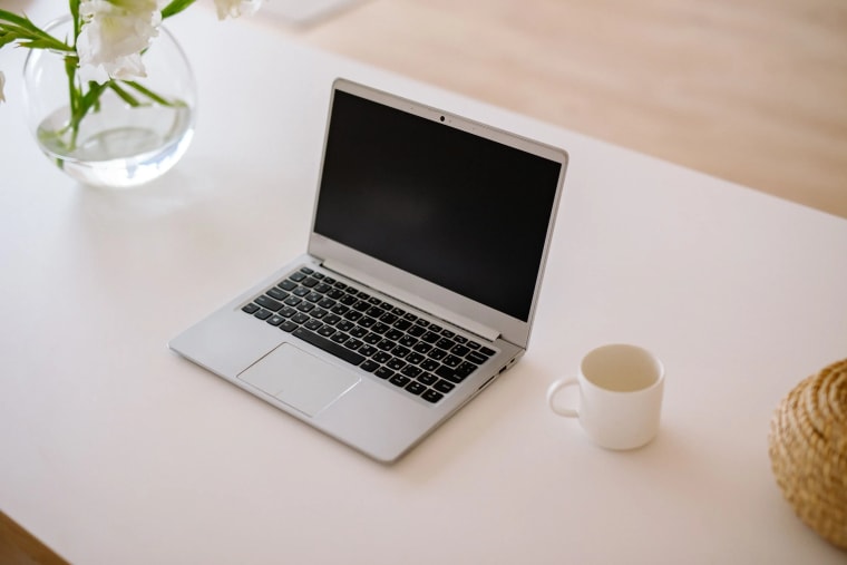 A laptop closed on a white table beside a glass vase of white flowers and a plain white mug.